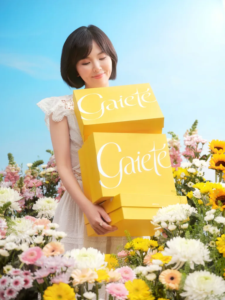 Woman smiling while holding yellow Gaiete product boxes against a bright sky and floral background.